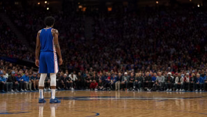 Player in a Philadelphia 76ers uniform standing alone on the court before a packed arena crowd